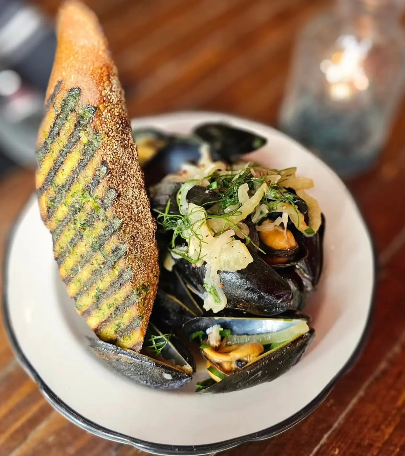 Steamed mussels in a white bowl topped with shaved fennel and herbs, served with a slice of grilled garlic bread