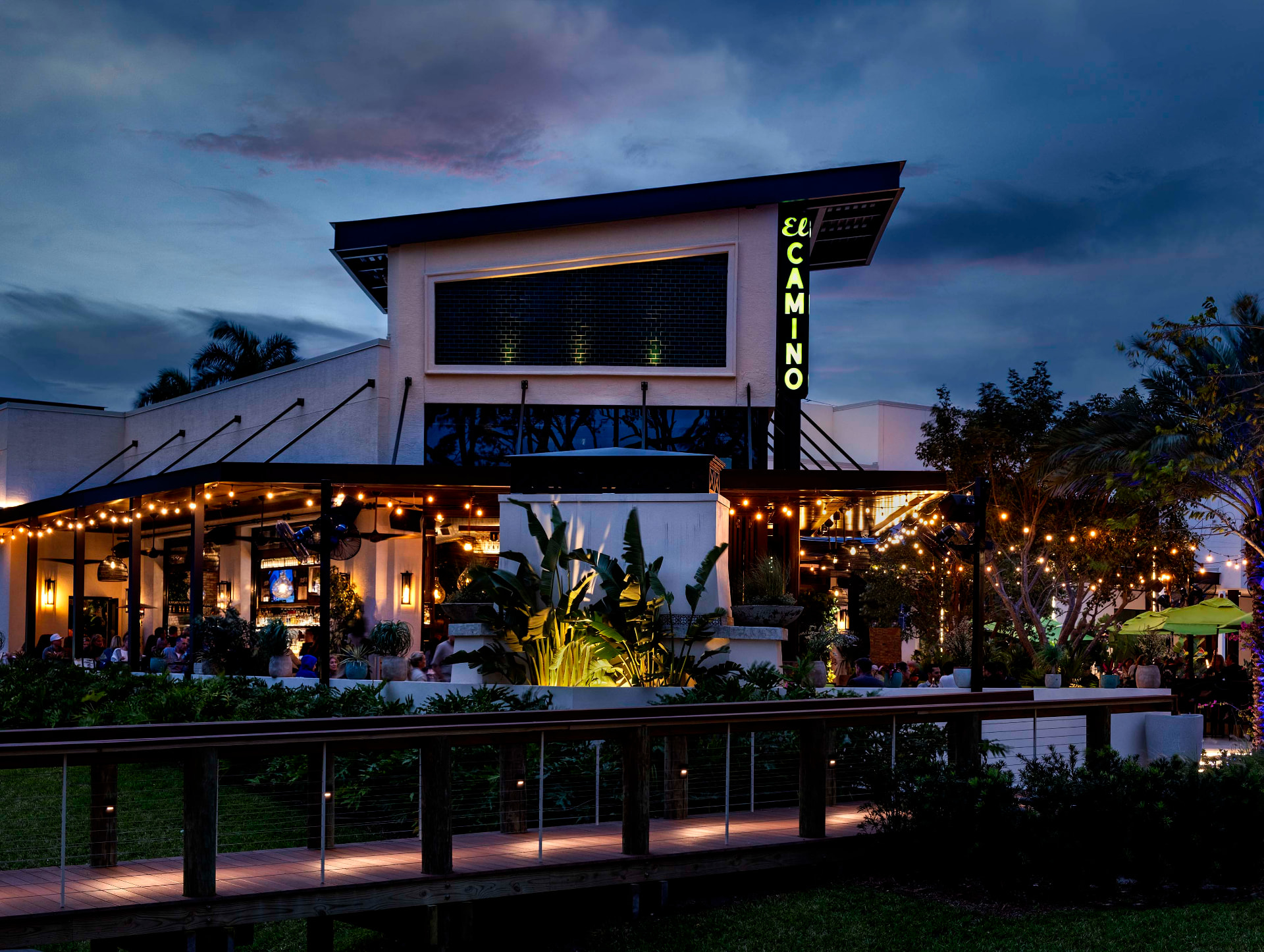 The exterior of El Camino at dusk — a glowing green neon sign on the building's corner tower, string lights over the patio, lush tropical plantings, and a boardwalk leading to the entrance