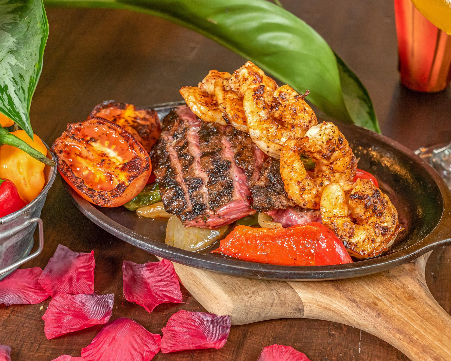 Grilled steak and chili-rubbed shrimp served sizzling in a cast-iron skillet with charred tomato and peppers, garnished with hibiscus petals