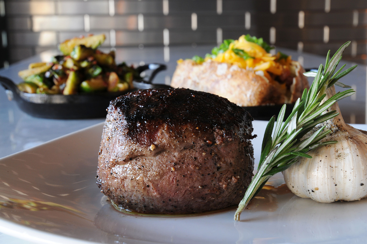 A char-grilled filet mignon at CUT 432 plated with a sprig of rosemary and roasted garlic, with a loaded baked potato and sautéed vegetables behind it